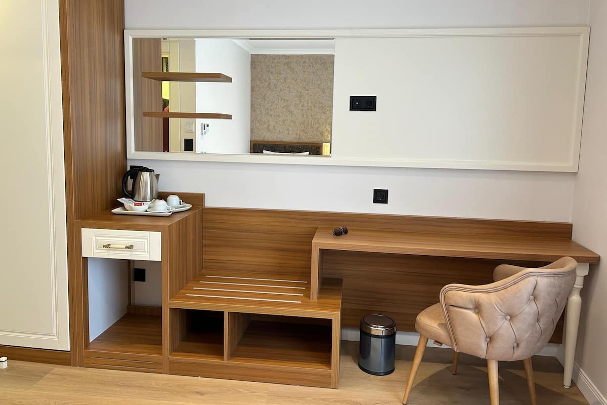 A compact hotel room with a wooden desk, a small seating area, a mirror, and a trash bin.