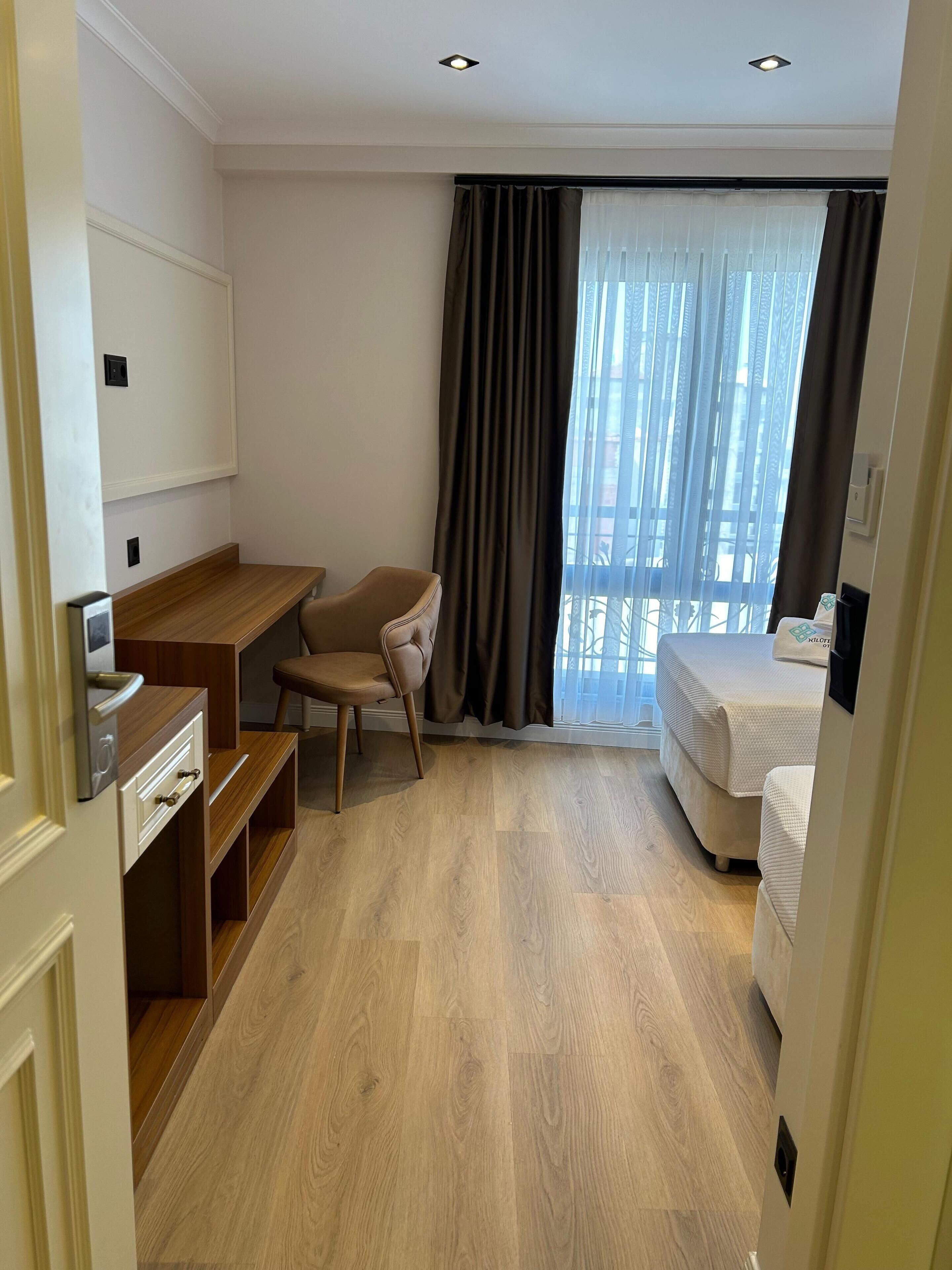 A hotel room with a wooden desk, a chair, a bed, and a large window with curtains.