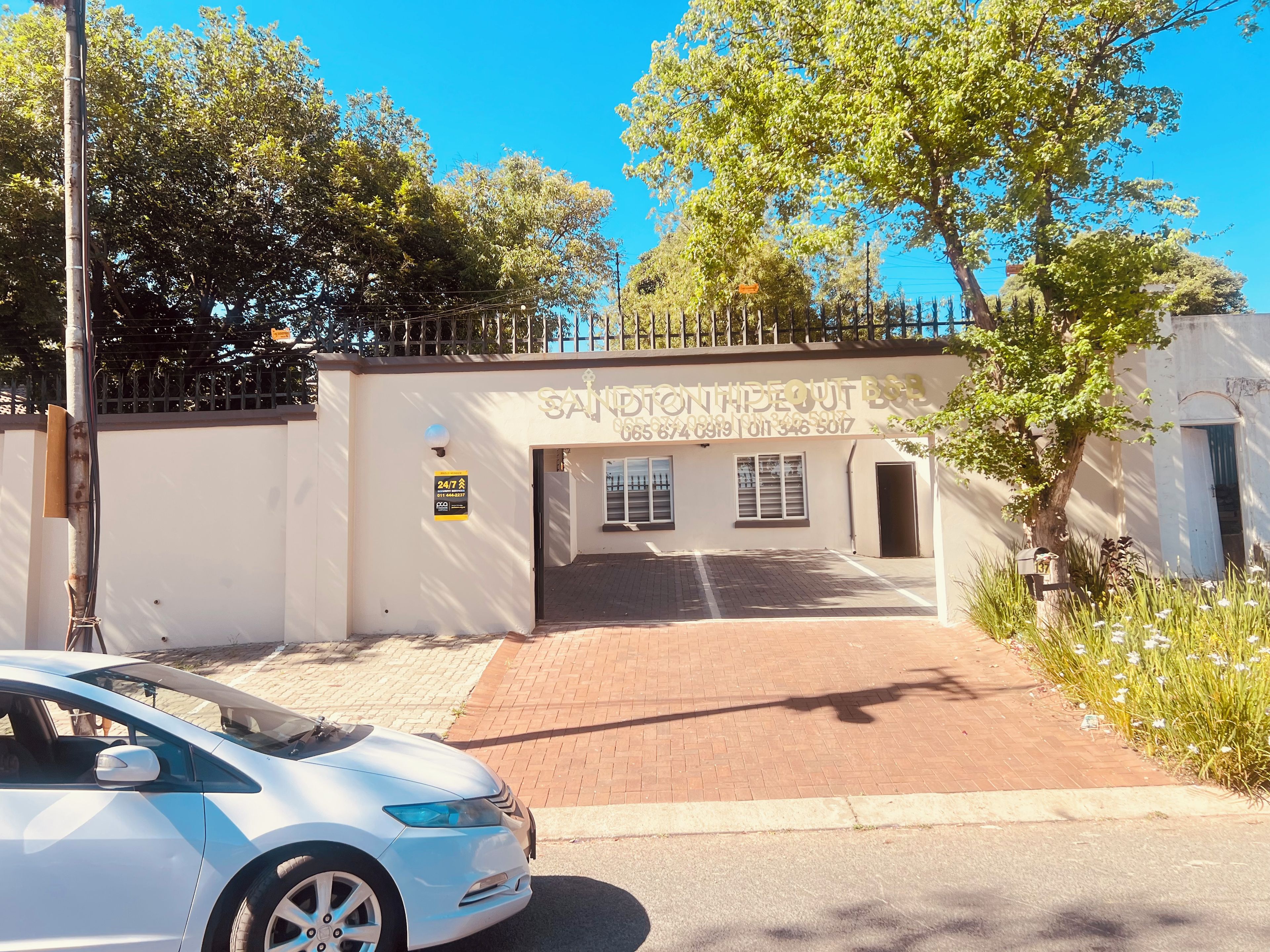 A white building with a sign that reads 'SANDTON HIDEOUT B&B' and a contact number. There is a paved entrance with a car parked in front.