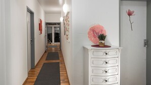 Een gang met een houten vloer, witte muren en een witte kast met lades. Er hangen afbeeldingen in lijsten aan de muur en op de kast staat een roze decoratie.