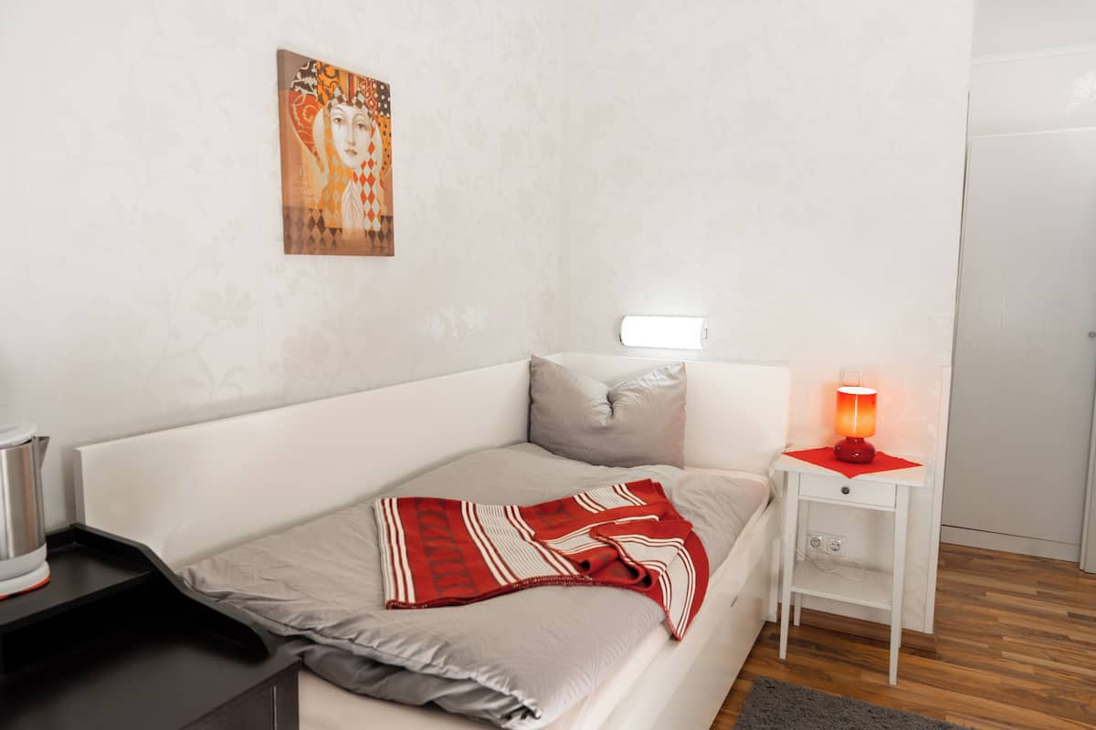 A neatly made bed with a red and white striped blanket, a white bedside table with a red lamp, and a wall-mounted artwork.