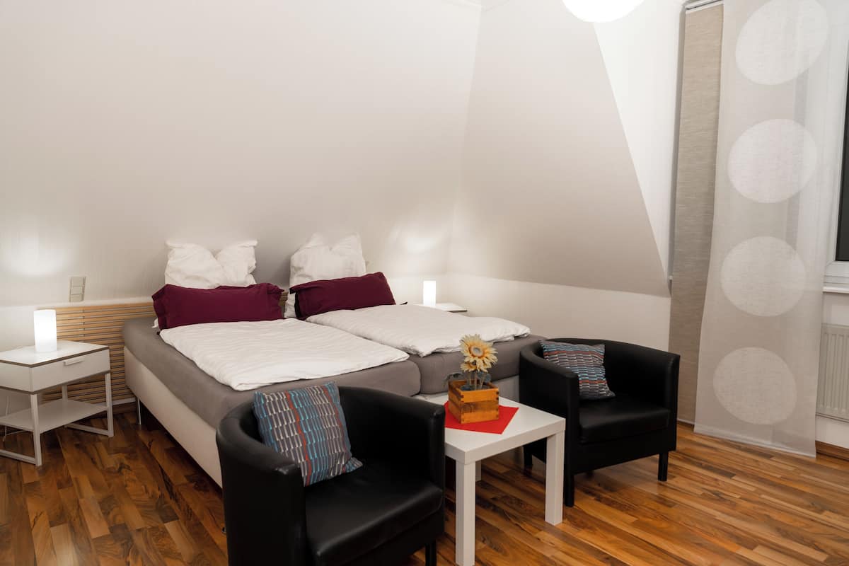 A modern bedroom with a bed, two nightstands, a small table, two chairs, and a large window with a curtain.