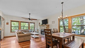 A spacious living room with a wooden floor, a ceiling fan, a fireplace, and large windows.