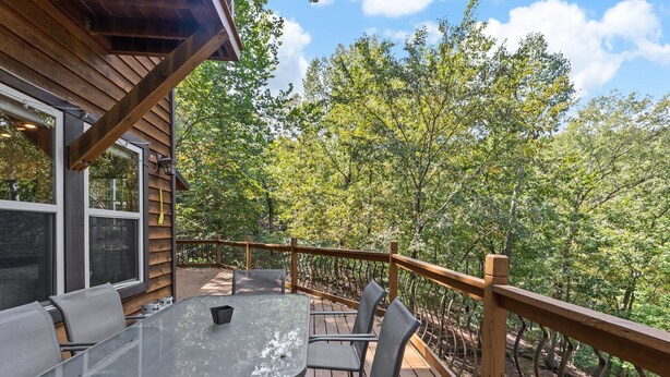A wooden deck with a glass table and chairs, overlooking a forested area.