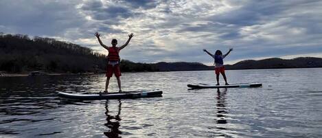 Två personer står på varsin ståpaddelbräda på en stilla sjö med en skogbeklädd strand och en molnig himmel.