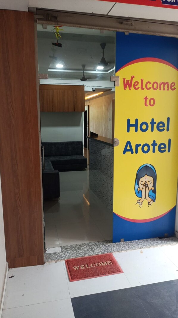 Entrada de un hotel con un cartel que dice 'Welcome to Hotel Arotel', un mostrador de recepción y un interior moderno con un sofá y ventilador de techo.