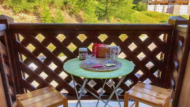 A small outdoor seating area with a green table and two wooden chairs, a woven placemat, a jar of Nutella, and a loaf of bread.