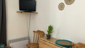 A living room with a television on a wall-mounted shelf, two wooden chairs, a small table with a plant, and a wooden cabinet.