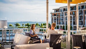 Una terraza con asientos al aire libre, vista al mar y un edificio con balcones al fondo.