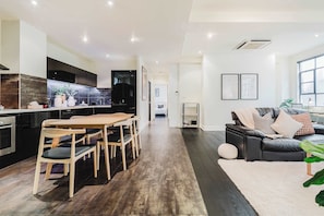 A modern kitchen with dark cabinetry, a dining area with wooden chairs, a black leather sofa, and a large window.
