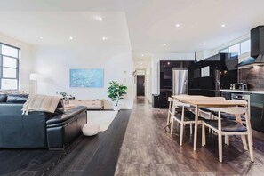 Uma sala de estar moderna com um sofá de couro escuro, uma mesa de jantar de madeira com cadeiras, um vaso com planta e um quadro na parede.