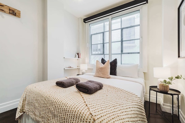 A hotel room with a large bed, a window, a bedside table with a lamp, and a small side table with a plant.