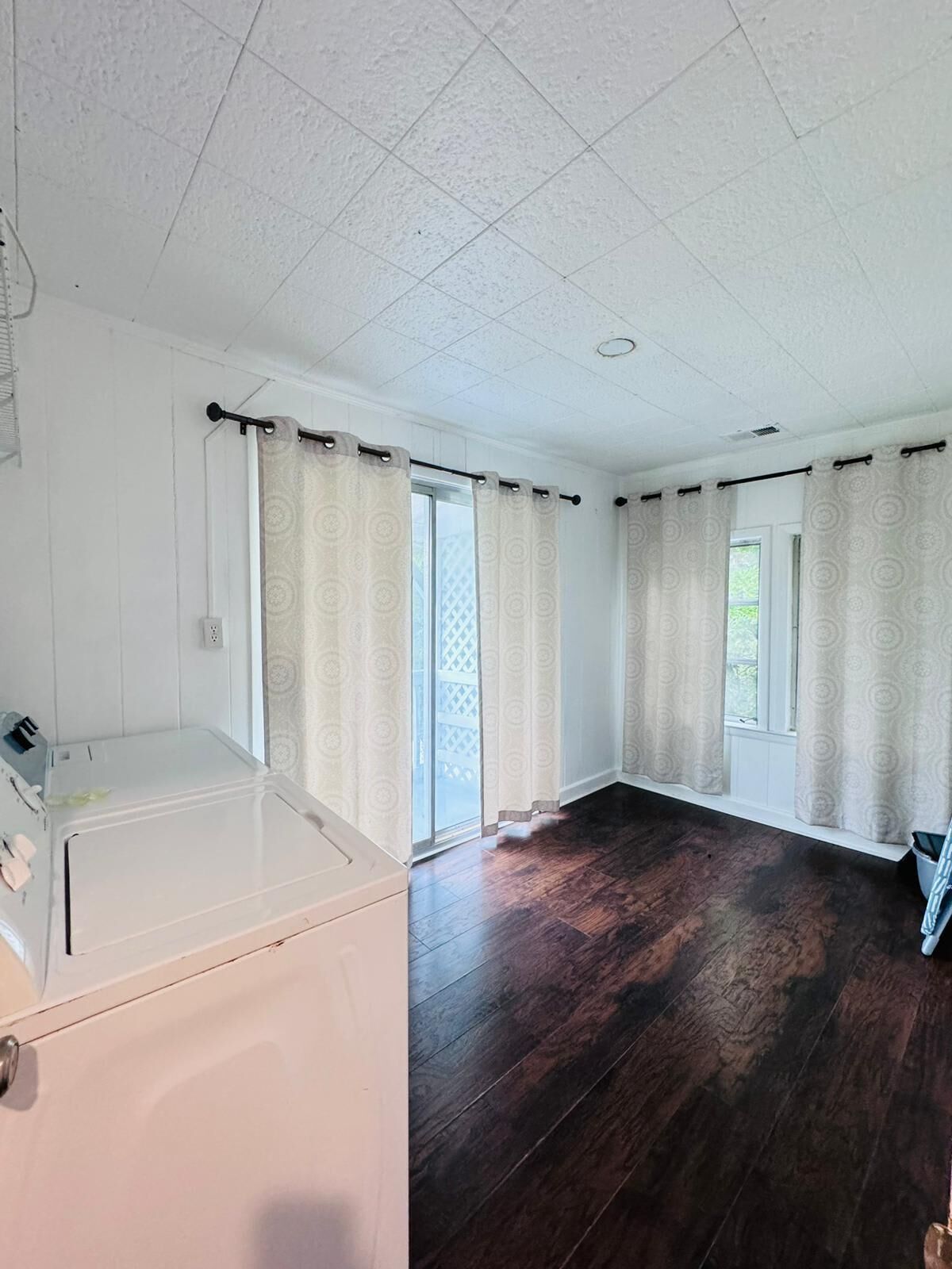Een kamer met een groot raam, een wasmachine en een donker houten vloer.