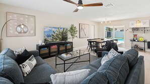 Un salon moderne avec un canapé, une table basse et un ventilateur de plafond. On aperçoit également un téléviseur, une bibliothèque et un coin repas à l’arrière-plan.