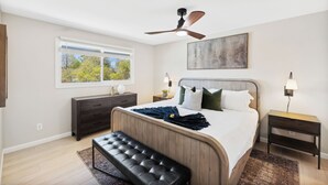 Une chambre à coucher comprenant un lit, un ventilateur de plafond, une table de chevet, une commode, un banc et une fenêtre donnant sur des arbres.