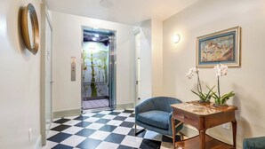 Un'accogliente hall d'hotel con pavimento a scacchi, una poltrona blu, un tavolino in legno con orchidee e un quadro appeso al muro.