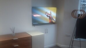 Un salon moderne avec un téléviseur à écran plat diffusant le film « Iron Man 2 », un meuble en bois, une petite table et une fenêtre équipée de stores.