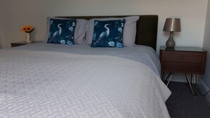 Un lit bien fait, recouvert d’une courtepointe matelassée, deux oreillers à motifs floraux bleus, une lampe de chevet et un petit vase avec des fleurs posé sur une table de chevet.