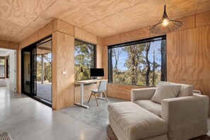 Ein modernes Interieur mit Holzboden, einem großen Fenster mit Blick auf Bäume, einem Schreibtisch mit Computer und einem Sofa.