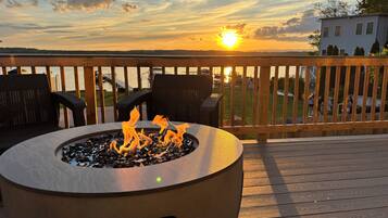 Una terraza con una chimenea y mobiliario exterior, con vista a un cuerpo de agua y una casa distante.
