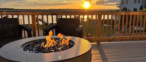 A deck with a fire pit and outdoor furniture, overlooking a body of water and a distant house.