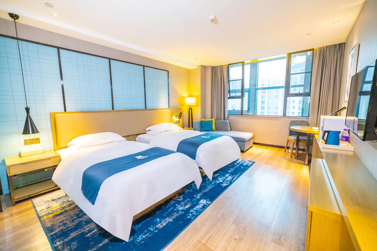 Quarto de hotel com duas camas, uma escrivaninha, uma cadeira e uma janela grande.