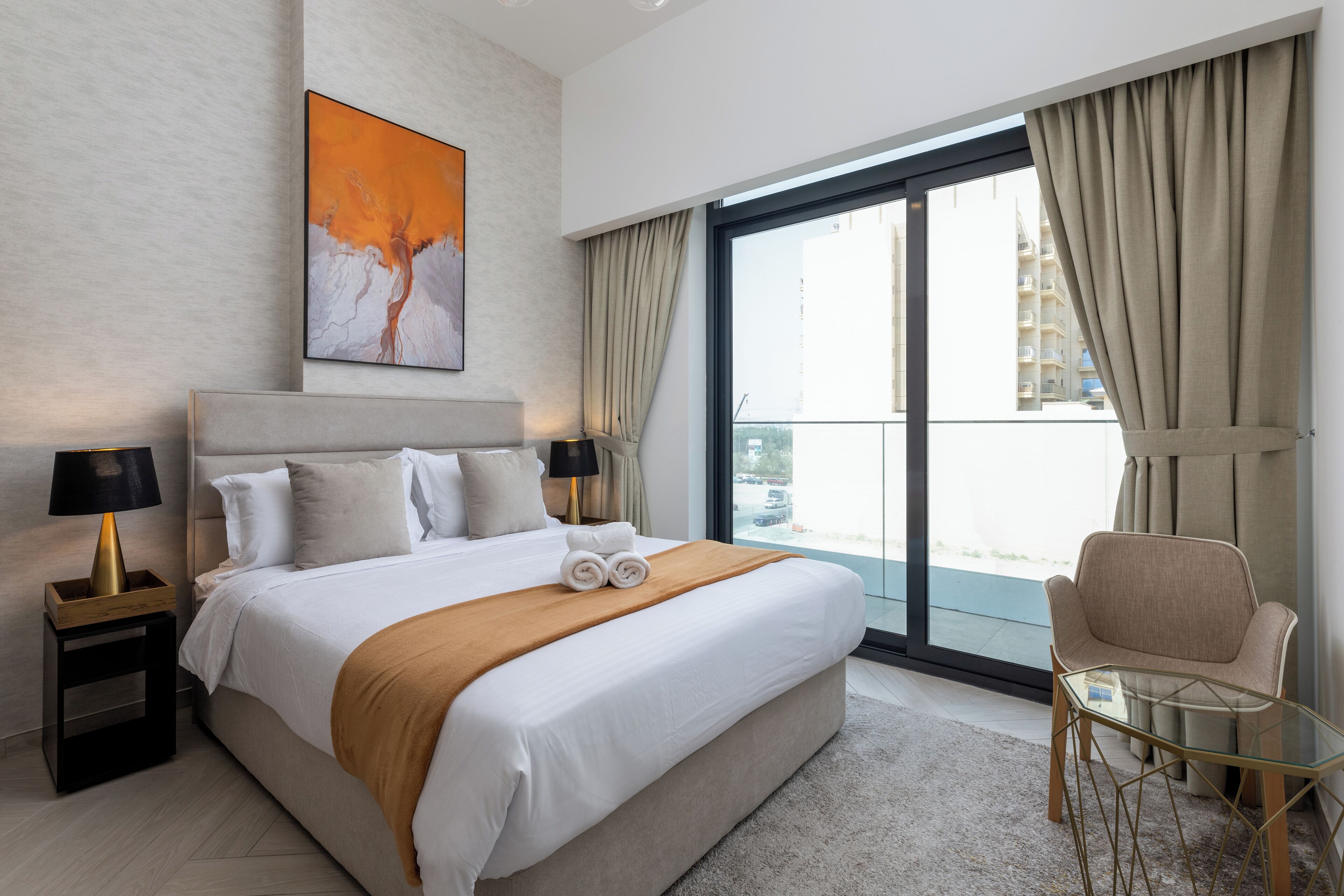 A modern hotel room with a large bed, a glass table, a chair, and a balcony with curtains.