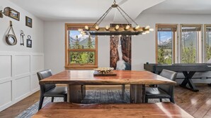 Un coin repas avec une table en bois, des chaises, un lustre et une vue sur les montagnes grâce à de grandes fenêtres.