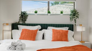 Un lit bien fait, avec une literie blanche et orange, une tête de lit verte et, par la fenêtre au-dessus, une vue sur un toit recouvert de tuiles.