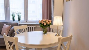 Ruokailutila, jossa on pyöreä pöytä, valkoiset tuolit, maljakko täynnä kukkia, lamppu ja tyynyillä varustettu sohva.
