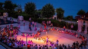 Una gran reunión al aire libre con gente sentada y de pie en un círculo, algunos en escalones y otros en el suelo, con un escenario en el centro.