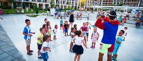A group of children and adults gathered in an outdoor plaza with a modern building in the background.