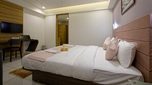 A modern hotel room with a neatly made bed, a desk with a chair, a flat-screen TV mounted on the wall, and a large mirror. - Hotel Aurellia (Ahmedabad)