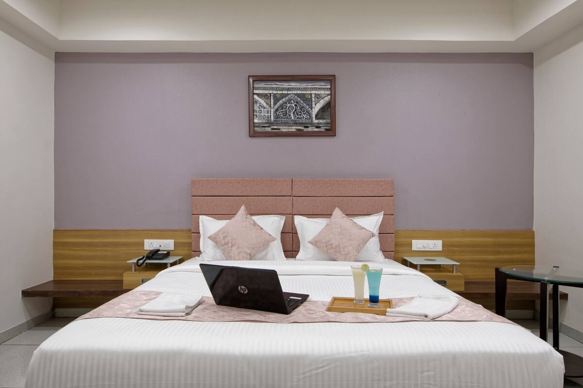 A hotel room with a bed, a laptop, a tray with drinks, and a framed artwork on the wall.