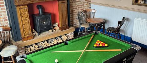 A pool table with a green felt surface, wooden chairs, a fireplace, and framed pictures on the wall.