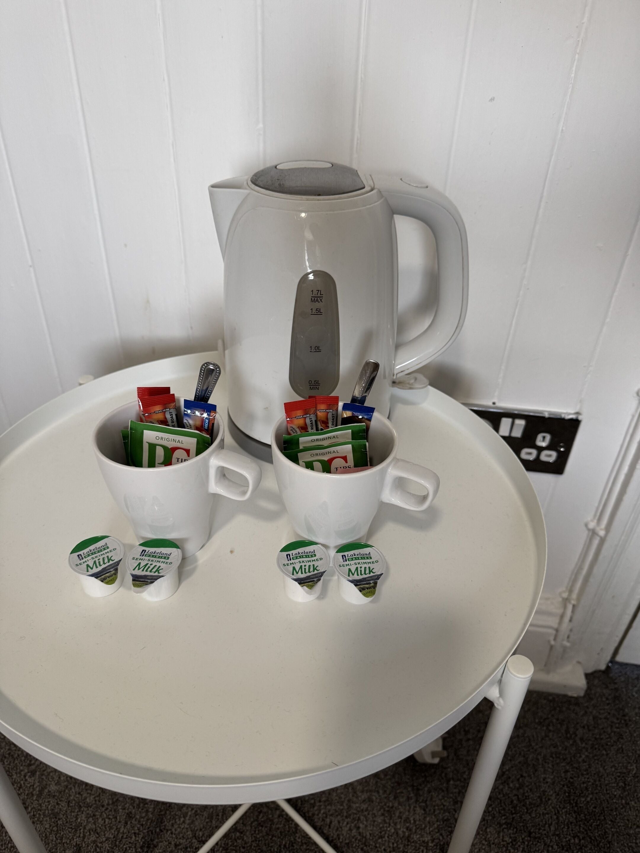 A white table with a white mug containing tea bags and a white mug with a tea kettle.