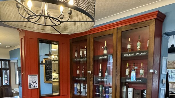 A cozy bar area with a leather couch, a wooden table, and a plaid chair. There are shelves with bottles labeled 'Champagnes,' 'Sparkling Wines,' and 'Wines of the Week.' A chandelier hangs above, and the walls are painted red. - Holystone Bar and Restaurant  (Newcastle-upon-Tyne)