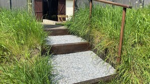 A rustic wooden staircase leading to a house with a tiled roof, surrounded by tall grass and gravel.