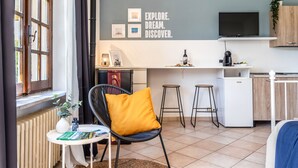 Eine moderne Küche mit weißer Arbeitsplatte, einem schwarzen Fernseher und einem kleinen Kühlschrank. Ein Stuhl mit gelber Sitzkissen und ein kleiner Tisch mit einer Zeitschrift.