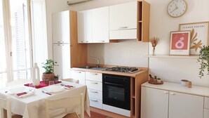 A compact kitchen with white cabinets, a stove, and a sink. There is a dining table with chairs and a clock on the wall. - Quick Home - Appartamento Narciso (Biella)