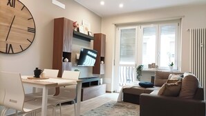 A modern living room with a dining area, featuring a large wall clock, a flat-screen TV, and a sofa. - Quick Home - Orchidea (Biella)
