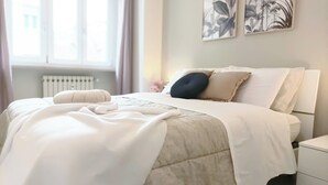 A neatly made bed with a white duvet, a dark pillow, and a beige throw blanket. There are two wall-mounted artworks depicting foliage. - Quick Home - Orchidea (Biella)