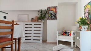 Een kamer met een houten stoel, een witte bank, een witte plank, een potplant en een kleurrijk schilderij aan de muur.