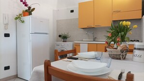 Een keuken met een witte koelkast, een wasmachine en een spoelbak. Er zijn houten keukenkastjes, een tafel met borden en bestek, en een vaas met bloemen.