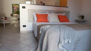 Een slaapkamer met een bed, witte muren, een betegelde vloer, een nachtkastje en een kamerplant, plus een schilderij.