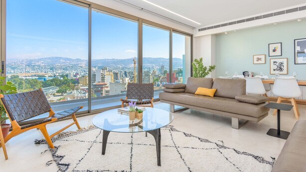Una sala de estar moderna con un ventanal grande, una mesa de centro de vidrio, un sofá y una zona de comedor con sillas.