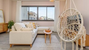 Un salon moderne avec un canapé, un fauteuil suspendu, une petite table et une grande fenêtre donnant sur des immeubles.