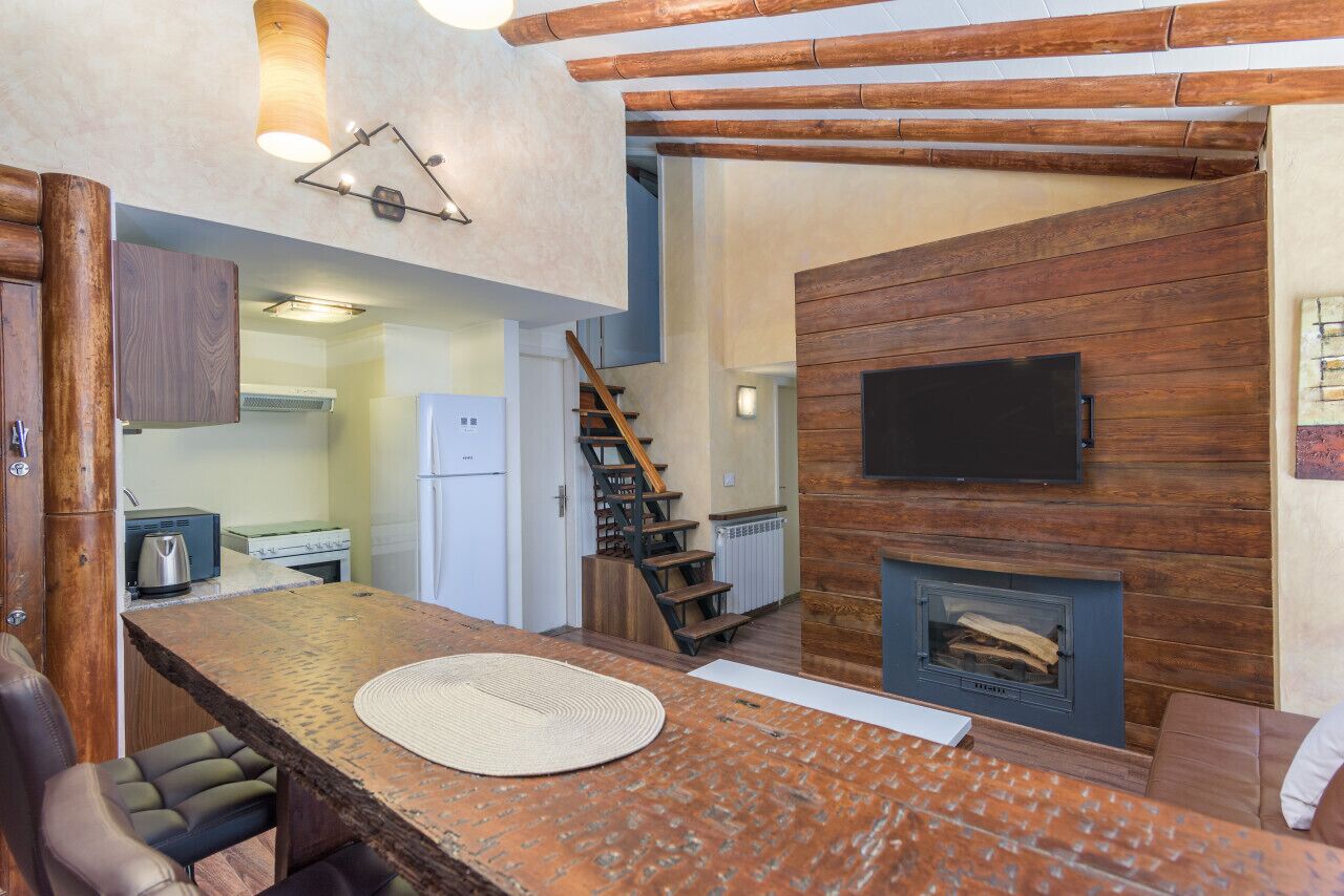 Una cocina moderna con estufa integrada, una zona de comedor con mesa y sillas de madera, y una chimenea sobre la cual está montado un televisor.