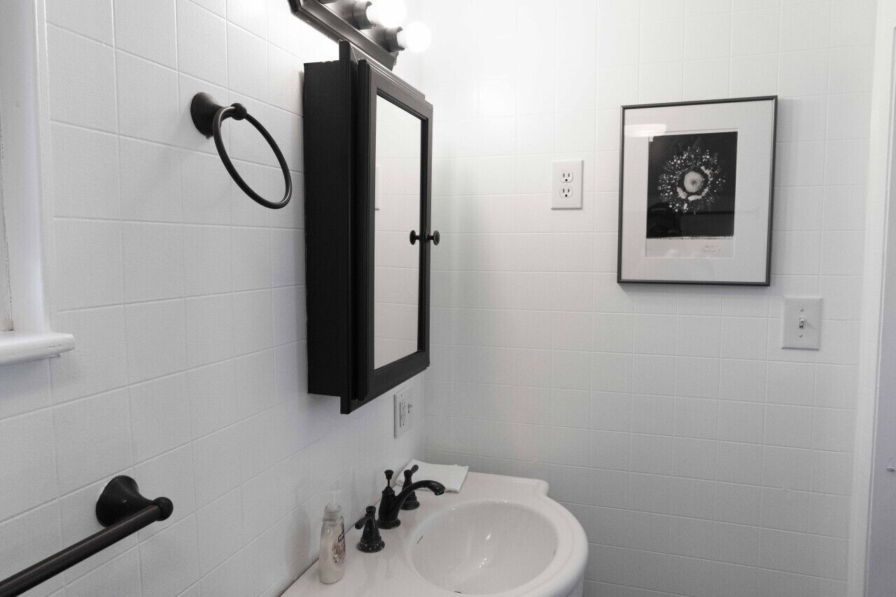A bathroom with white tiled walls, a black-framed mirror, a white sink, and a framed picture on the wall.