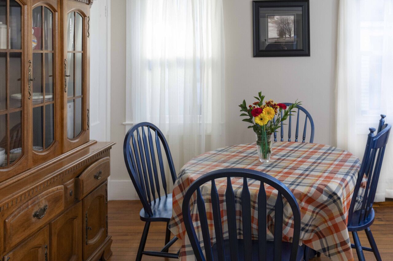 Un comedor con mantel escocés, sillas azules, un armario de madera y un jarrón de flores.